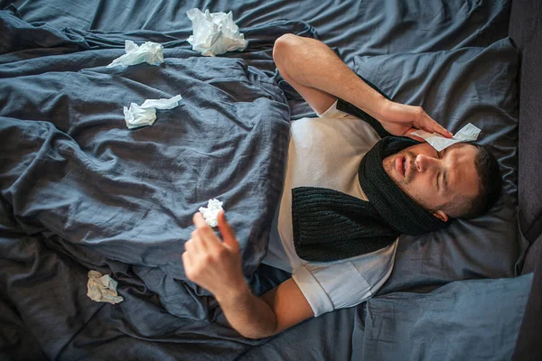 Foto do jovem deitado na cama e sofrendo de dor de cabeça e corrimento nasal. Ele sente-se mal e exausto. Jovem tem guardanapo seco na cabeça e encolhendo. Há muitos guardanapos na cama. . — Fotografia de Stock