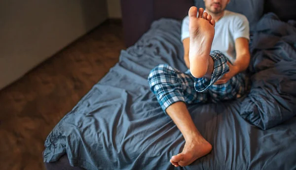 O jovem senta-se na cama e mostra o pé na câmera. Ele segura a perna com as duas mãos. O tipo está no quarto. Vista para corte . — Fotografia de Stock