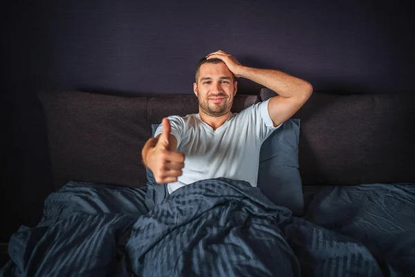 Rolig ung man sitter på sängen och ler. Han visar stor tumme upp å ena sidan och håll en annan en på huvudet. Killen är nöjd och glad. — Stockfoto