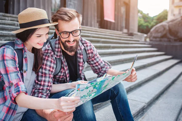 Young man and woman sit on steps outside. They hold map together. Tourists smile. They look at map. Travellers have rest. — Stock Photo, Image