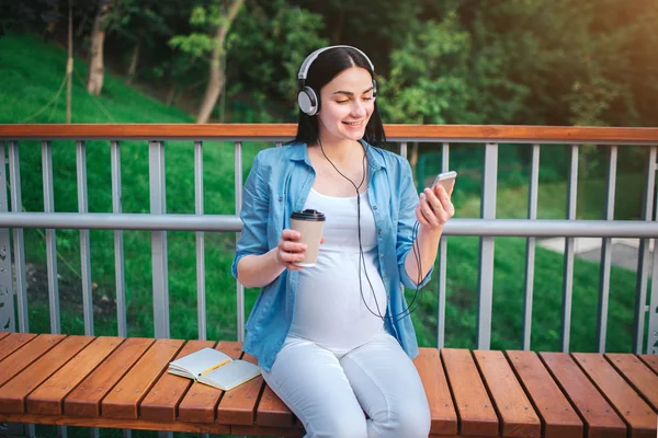 Portrait d'une heureuse chevelure noire et fière femme enceinte dans une ville en arrière-plan. Elle est assise sur un banc de la ville. La future mère écoute de la musique dans le parc avec un enfant à naître — Photo