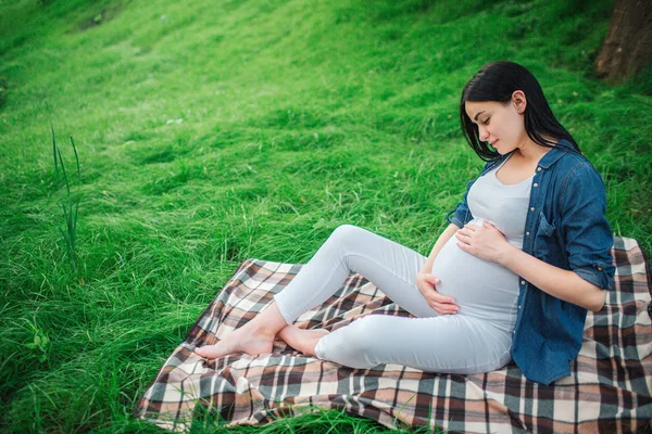 Portrait of a happy black hair and proud pregnant woman in a city in the park . Photo of female model touching her belly with hands. The female model is sitting on grass . — Stock Photo, Image