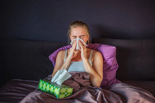 Mujer joven enferma y enferma estornudando en una servilleta blanca. Se sienta en la cama y se cubre con manta de seda. Mujer joven tiene caja verde de pañuelos en la cama . — Foto de Stock