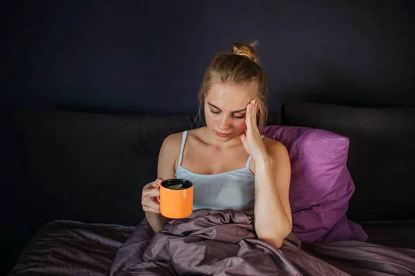 Poważne i martwi się młoda kobieta siedzi na łóżku. Ona trzyma kubek herbaty z cytryną i trzyma rękę na czole. Ona ma ból głowy. Młoda kobieta jest cierpieć. — Zdjęcie stockowe
