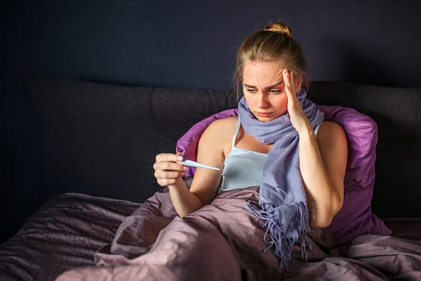 Bardzo chory młoda kobieta siedzi na łóżku i wygląda na termometr, który trzyma w ręku. Inny jeden jest na czole. Bloden ma szalik wokół szyi. — Zdjęcie stockowe