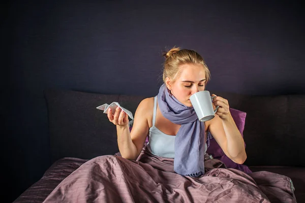 Unga sjuka kvinnan sitter på sängen och drycker från vita cup. Hon håller torr vävnad i en annan hand. Hennes ben är täckta med filt. — Stockfoto