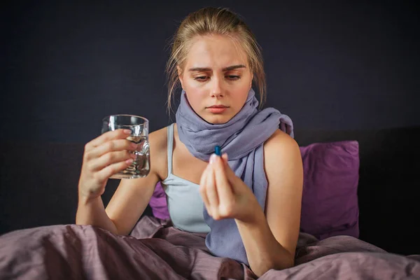 Сконцентрированная молодая женщина сидит на кровати и смотрит на лекарство, которое держит пальцами. В другой руке стакан воды. . — стоковое фото