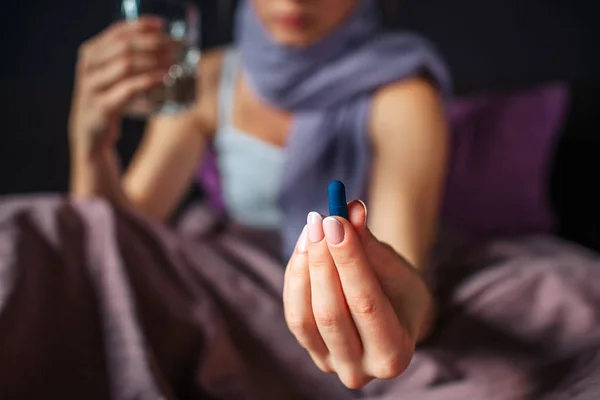 Vue coupée de la jeune femme assise sur le lit et montre la médecine à la main. C'est une capsule bleue. Il y a un verre d'eau dans une autre main. Son cou est couvert d'écharpe . — Photo