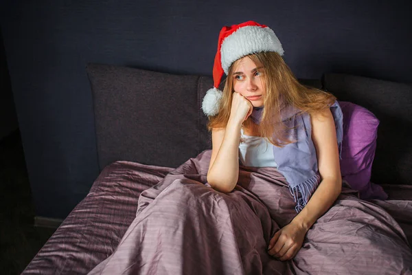 Chory młoda kobieta siedzi na łóżku i wygląda prosto. Ona się nudzi. Kobieta trzyma ręce pod brodą. Ona ma Boże Narodzenie kapelusz na głowę. — Zdjęcie stockowe