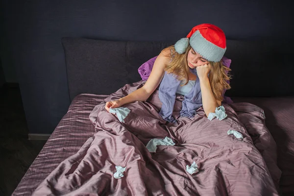 Znudzony i chory młoda kobieta w Boże Narodzenie kapelusz siedząc na łóżku i trzymając w ręku tkanki. Istnieje inny zużytych serwetek, leżąc na łóżku. Ona się nudzi. — Zdjęcie stockowe