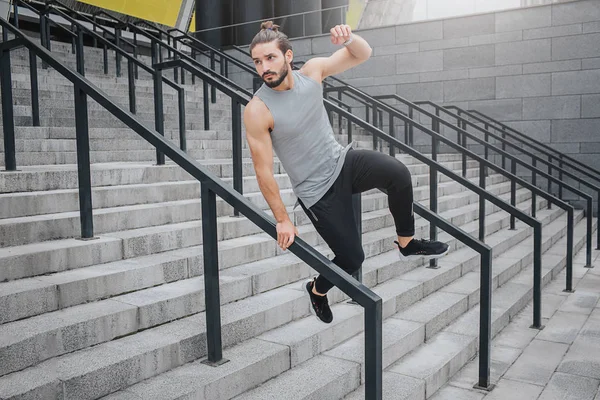 Young man is in motion. He holds one hand on barrier and jumps over it. He looks straight. Young man jumps on stairs. He is confident and experienced.