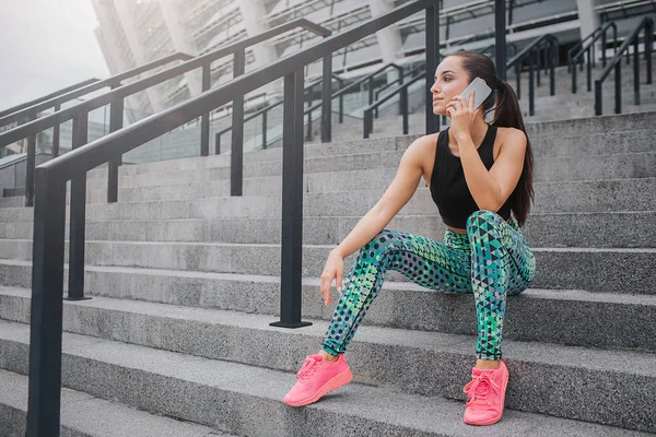 Mujer joven positiva y alegre se sienta en los pasos y habla por teléfono. Descansa después de entrenar duro. La joven sonríe un poco. Parece confiada. . —  Fotos de Stock