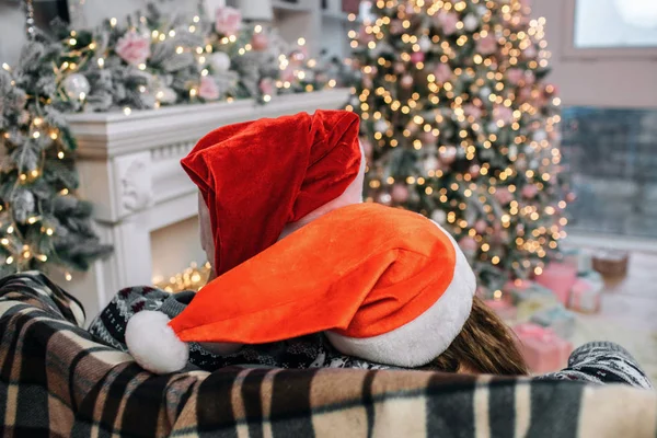 Deux chapeaux de Noël sont sur la tête des gens. Elle se penche vers lui. Ils sont assis sur le canapé. Les gens sont dans la salle de Noël décorée . — Photo