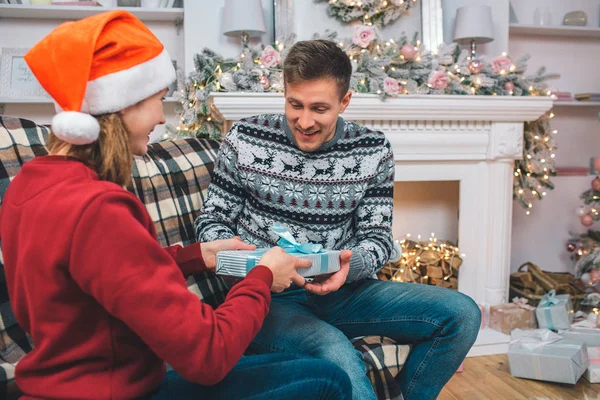 Verblüffter junger Mann sitzt vor Frau und blickt in die Gegenwart. Er hat es von der Frau. Er ist glücklich. junge Frau mit rotem Hut hält Geschenk. — Stockfoto