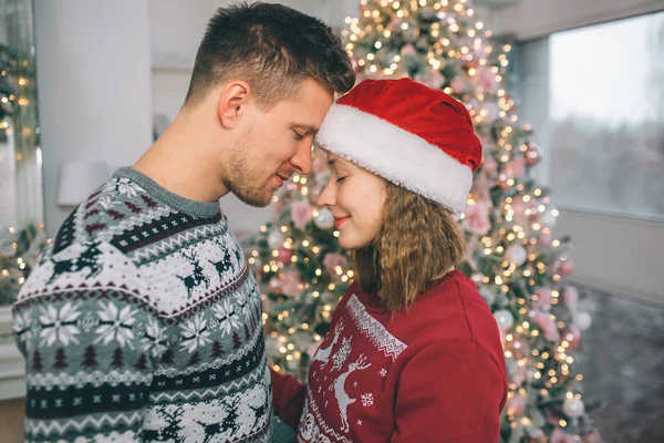 Pareja pacífica y atractiva que mantiene los ojos cerrados y se toca la frente. Se paran frente al árbol de Navidad en la habitación decorada . — Foto de Stock