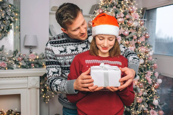 Junger Mann steht hinter Frau. Sie halten Schachtel mit Geschenk zusammen. schaut er sie an. , blickt sie in die Gegenwart. sie befinden sich im dekorierten Raum. — Stockfoto