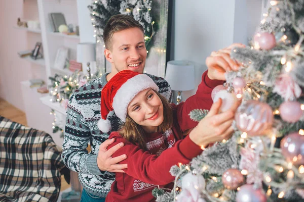Junge Frau schmückt Weihnachtsbaum. sie berührt Spielzeug darauf. junger Mann steht hinter ihr und umarmt sie. sie lächeln. — Stockfoto