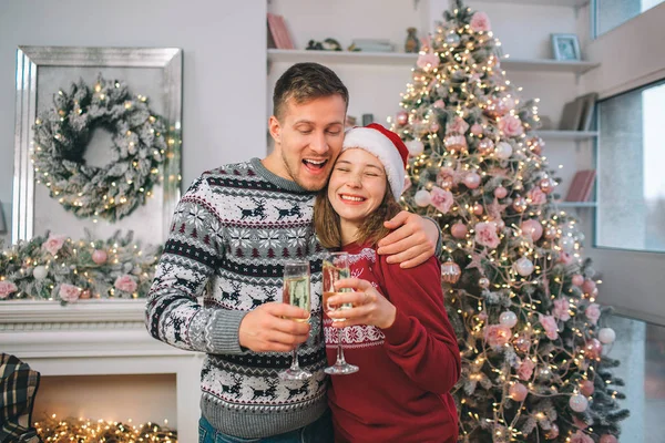 Positives Paar steht zusammen. junger Mann umarmt Frau. Sie hält die Augen geschlossen und lächelt. Jeder von ihnen hat ein Glas Sekt. Menschen stehen in dekoriertem Raum. — Stockfoto