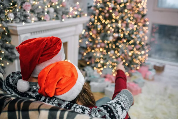 Photo de l'homme et de la femme assis sur le canapé. Ils portent des chapeaux de Noël rouges. Elle pointe du doigt l'arbre de Noël. Il l'embrasse. Les gens sont dans la chambre décorée . — Photo