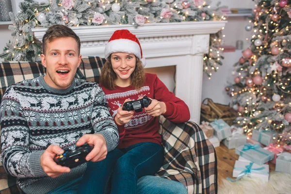 Zwei Personen sitzen und spielen. thay halten Gamepads in den Händen. Junge Männer und Frauen sind begeistert. Sie drücken Knöpfe und spielen aufgeregt im dekorierten Raum. — Stockfoto