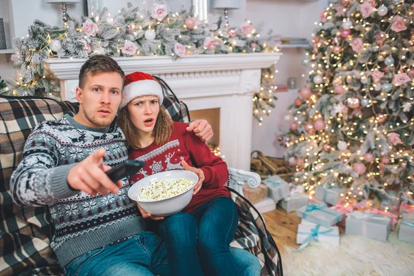 Bild eines jungen Mannes und einer jungen Frau, die sitzen und sich umarmen. Sie sind erstaunt. Menschen, die geradeaus schauen. Sie hält eine Tüte Popcorn in der Hand. er hat eine Fernbedienung. — Stockfoto