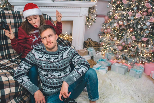 Die verspielte junge Frau sitzt auf dem Sofa und zeigt Zunge. Sie schaut ihn an. er sitzt aufgebracht auf dem Boden. Mann verlor Spiel. Frau ist Gewinnerin. Sie ist glücklich. — Stockfoto