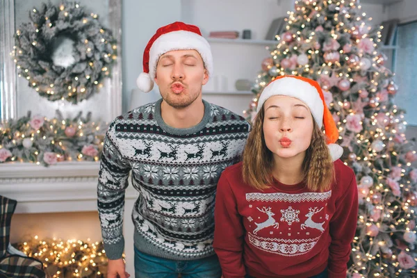 Divertido cuadro de hombre y mujer jóvenes se levanta y posa. Mantienen los ojos cerrados. Los labios de la gente están en forma de beso. Hay árbol de Navidad y chimenea detrás de ellos . — Foto de Stock
