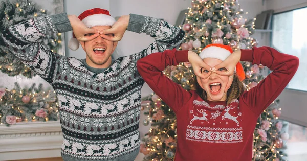 Les adultes drôles se tiennent debout et tiennent les doigts autour des yeux. Ils sourient et gardent la bouche ouverte. Les gens portent des pulls de Noël et des chapeaux rouges . — Photo