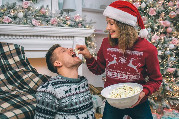 Schöne Bild der jungen Frau steht und füttert ihren Freund. Sie hält ihm ein Stück Popcorn über den Mund. Mann hält Mund offen. Junger Mann sitzt auf dem Boden. — Stockfoto
