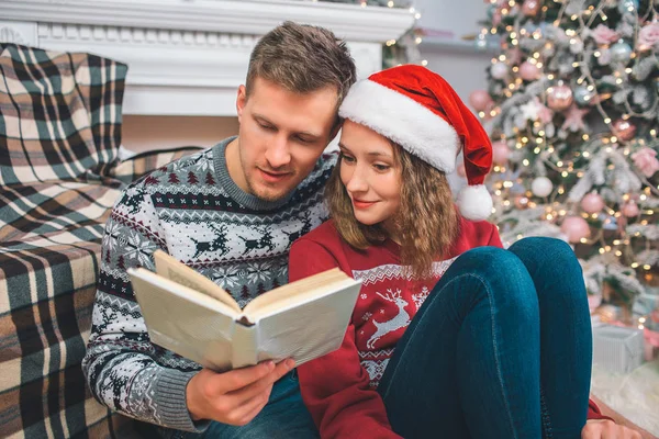 Picture of young man and woman sitting together on floor. They read book. He holds it in hands. Woman smiles a bit. They are in decorated room. — Stock Photo, Image