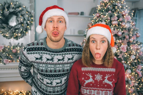 Un jeune homme et une jeune femme en tenue de fête regardent la caméra. Ils se tiennent ensemble et montrent leur langue. Il garde les mains derrière lui. Ils portent des chapeaux de Noël . — Photo