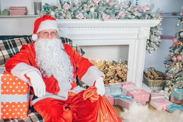 Photo de l'homme en costume santa claus assis et pose sur la caméra. Il tient une main sur la boîte cadeau. Il ya cheminée avec des chaussettes et des cadeaux derrière lui . — Photo