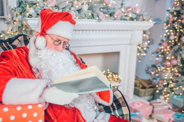 Calme et paisible homme en costume de Père Noël est assis et lire le livre à travers des lunettes. Il fait attention. Un homme seul dans la chambre. Il a un look vénitien . — Photo