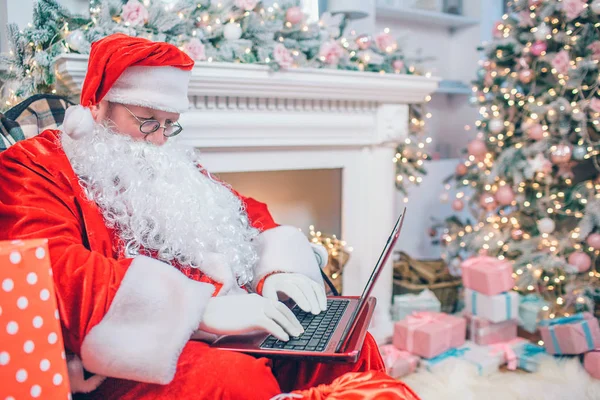 El moderno Santa Claus se encuentra junto a la chimenea y el árbol de Navidad. Él sostiene el ordenador portátil en la siesta y trabaja. Guy está concentrado . —  Fotos de Stock