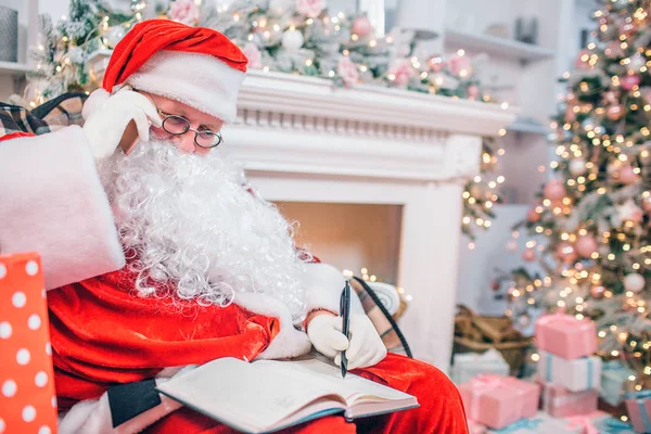 L'homme concentré et réfléchi dans le costume du Père Noël est assis à la cheminée et à l'arbre de Noël. Il tient le téléphone t oreille et les blancs vers le bas dans le cahier . — Photo