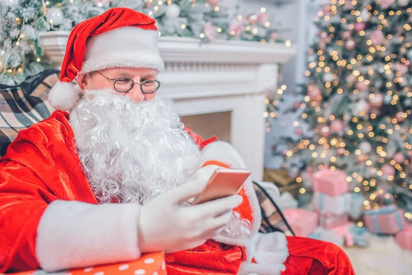 Père Noël occupé s'assoit et regarde le téléphone qu'il tient dans ses mains. L'homme est seul dans la salle des fêtes. Il est assis à la cheminée et au sapin de Noël . — Photo