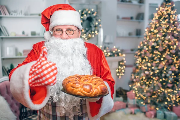 Szczęśliwy człowiek w Santa Claus ubrania posiada pie i pokazuje wielki kciuk. Facet wygląda na aparat. On jest w uroczysty pokoju sam. — Zdjęcie stockowe