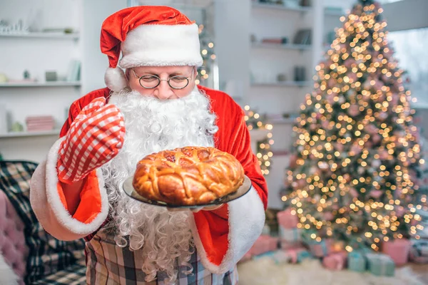 Tarte fraîche et savoureuse est entre les mains du Père Noël. Il le regarde et sent. Il y a un sapin derrière lui. . — Photo