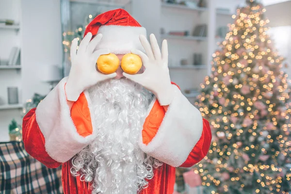 Père Noël ludique et drôle couvre les yeux avec deux mandarines. Il porte des vêtements de Noël . — Photo