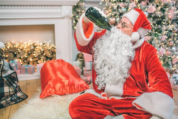 Santa Claus siedzi na podłodze i pije alkohol z butelki. On jest pijany. Jego czerwone worek jest kominek. Człowiek sam w pokoju. — Zdjęcie stockowe