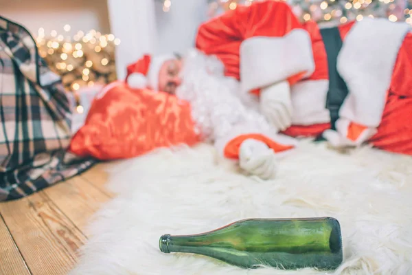 Obraz pijany mężczyzna w Santa Claus ubrania spanie na podłodze. On ma czerwony worek pod głową. Kamera koncentruje się na zielone butelki na piętrze. — Zdjęcie stockowe