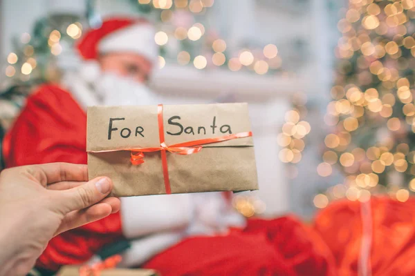 Imagen de la mano sosteniendo carta para Santa. Está cubierto con cinta roja. Hay Papá Noel sentado y sosteniendo cartas. Los lee. . — Foto de Stock