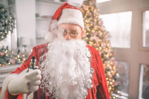 Homme détendu dans le costume du Père Noël se tient debout et tient vape dans la main. La fumée sort de sa bouche. Il est détendu. . — Photo