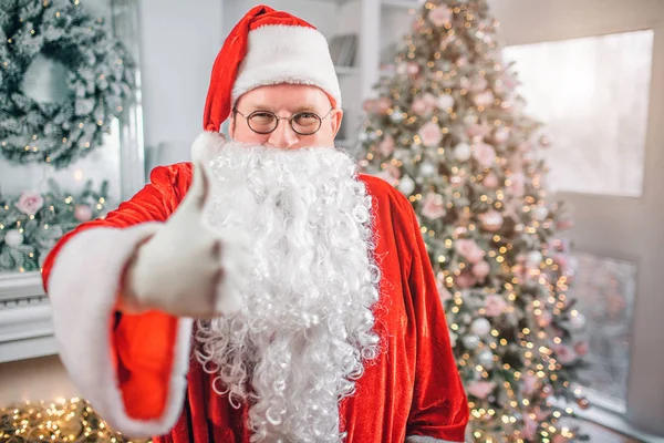 Positive and happy Santa Claus looks on camera and holds big thumb up. He is in room alone. Man stands in front of fireplace and Christmas tree. — Stock Photo, Image