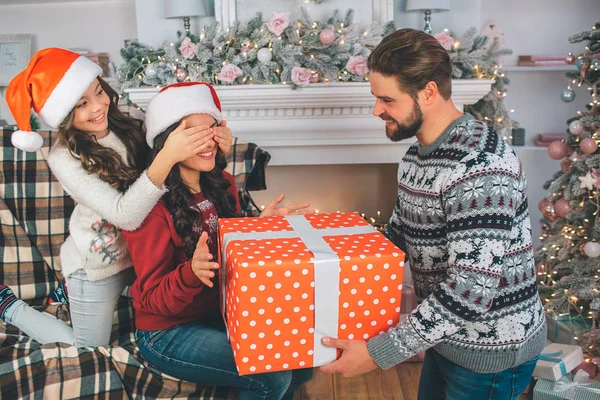 Joven hombre y mujer sosteniendo una gran caja de regalo. La niña cierra los ojos a sus madres. El hombre mira a su esposa. La familia viste ropa festiva . — Foto de Stock