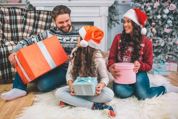 Fröhliche und positive Familie sitzt auf dem Boden und lächelt. schauen sie einander an. Vater hält große Kiste mit Geschenken. Die junge Frau hat ein rosafarbenes, das Mädchen eine blaue Schachtel in der Hand. Sie sehen festlich aus. — Stockfoto