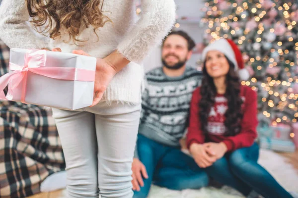 Bonita foto de chica stnds delante de los padres. Ella tiene un regalo. La chica lo esconde detrás de sí misma. Los padres se sientan en el suelo y lo miran. Sonríen . — Foto de Stock