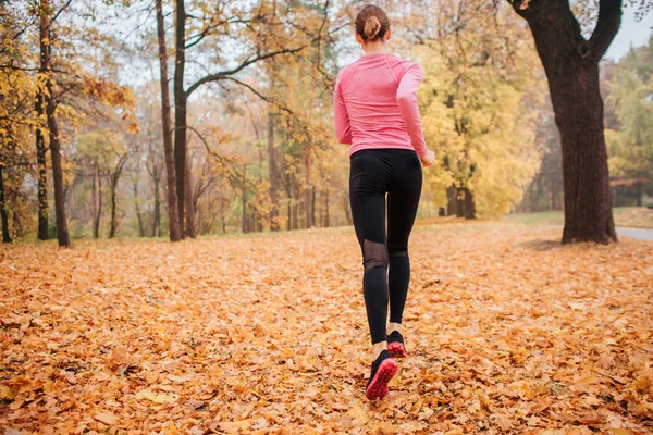 Picture of young woman running in park. It is chilly weather outside. Orange leaves lying on ground. Young woman jogging. She run alone.