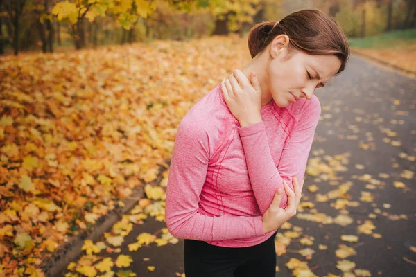 Sad and upset young woman suffer from pain in neck and elbow area. She keep seyes closed. Woman is in park alone. — Stock Photo, Image