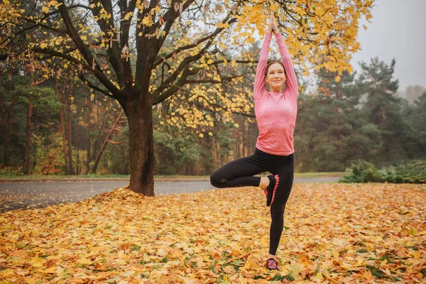 Calm, peaceful and positive young woman stands in asana position and look straight. She holds hands up. Woman is in autumn park alone.